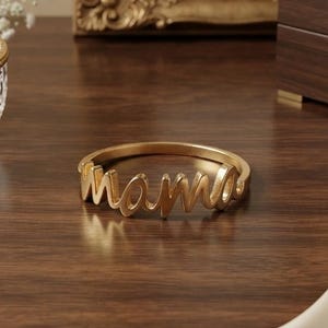 Könnte beinhalten: Ein goldfarbener Ring mit dem Wort mama in einer kursiven Schreibschrift. Der Ring besteht aus einem polierten Metallband, das die Buchstaben des Wortes formt und ein durchgehendes Design bildet, das flach auf der Oberfläche liegt.