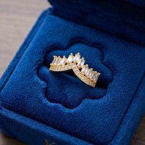 May include: A gold chevron-shaped ring featuring a row of baguette-cut clear stones along the top edge and a band of small round pavé-set stones underneath, resting inside a deep blue velvet ring box with a scalloped interior lining.