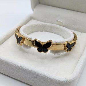 Puede incluir: Una pulsera rígida articulada de tono dorado con tres motivos de mariposa espaciados uniformemente. Cada mariposa tiene una incrustación negra lisa rodeada por un borde dorado con cuentas. La pulsera tiene un acabado pulido y diseño simple.
