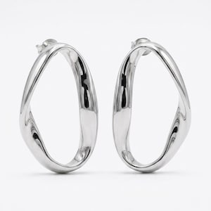 Op de afbeelding: Een paar zilverkleurige oorbellen met een onregelmatige, open ovale vorm en een glad, reflecterend metalen oppervlak. Elke oorbel heeft een gedraaid, vloeiend ontwerp dat een doorlopende lus vormt, bevestigd met een standaard stekersluiting.