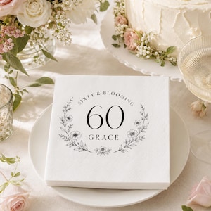 May include: A square white paper napkin featuring a black printed design of a floral wreath surrounding the number 60 and the text Sixty & Blooming Grace. The napkin is centered on a white plate, showcasing a clean and simple aesthetic.