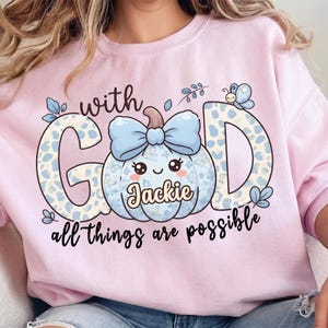 Op de afbeelding: Een lichtroze sweater met ronde hals en een grafische print met de tekst "with GOD all things are possible". Het ontwerp bevat een lichtblauwe pompoen met een strik, een vlinder en de naam "Jackie" geschreven in cursief schrift.