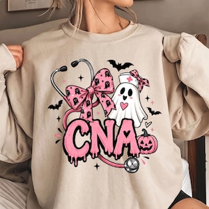 May include: A beige crewneck sweatshirt featuring a graphic print of a pink stethoscope, a ghost wearing a nurse cap with a pink cross, a pink pumpkin, and a pink bow with ghost patterns. The text CNA is printed in large, pink, dripping letters.