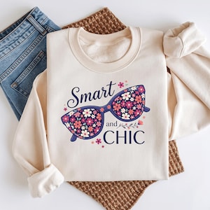 Op de afbeelding: Een crèmekleurig sweatshirt met ronde hals en een afbeelding van een donkerblauwe cat-eye bril gevuld met een roze en wit bloemenpatroon. De tekst "Smart and CHIC" is in donkerblauwe letters rond het brilontwerp gedrukt.