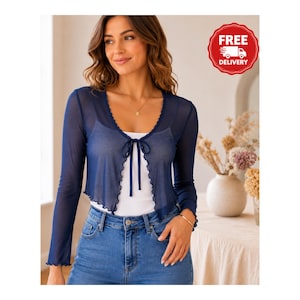 May include: A navy blue sheer long-sleeve bolero shrug featuring a front tie closure and lettuce-edge trim along the neckline and hem. The garment is worn over a plain white camisole and paired with blue denim jeans, showcasing a lightweight mesh fabric.