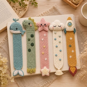 May include: Five crocheted bookmarks featuring a blue whale, a green dinosaur, a pink star with embroidered sparkles, a white cloud with blue dots, and a yellow rocket ship with a flame base, all arranged vertically on a white notebook.