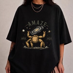 May include: A black short-sleeve t-shirt featuring a graphic print of a tan mechanical crab-like creature making peace signs. Above the creature is a spiral galaxy, planets, and the text "AMAZE" in a serif font. The design includes small stars.