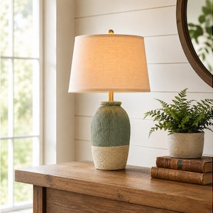 May include: A table lamp featuring a textured, two-toned ceramic base with a sage green upper section and a speckled cream lower section. The lamp is topped with a tapered, off-white fabric drum shade and a small gold-toned finial at the top.