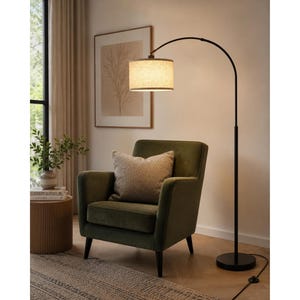 May include: A tall, black metal arc floor lamp with a cylindrical, light-colored fabric drum shade. The lamp features a slender, curved arm extending from a circular base, providing downward illumination over an olive green upholstered armchair.