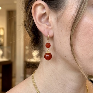 Può includere: Un orecchino pendente dorato con due perle rotonde rosso scuro di dimensioni diverse. La perla più piccola si trova sopra quella più grande, decorata con una coppetta in metallo dorato. L'orecchino è fissato a un semplice gancio dorato.