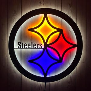 May include: A circular wall sign featuring the Steelers logo with the word Steelers in black lettering. The design includes three four-pointed star shapes illuminated from behind in yellow, red, and blue light, surrounded by a black circular frame.