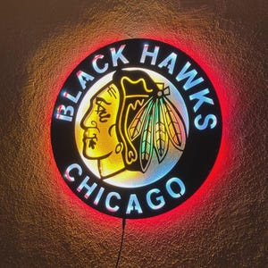 Può includere: Un'insegna da parete circolare con il logo dei Chicago Black Hawks che mostra il profilo di una persona con un copricapo. Il design è illuminato con un effetto luminoso e mostra il testo BLACK HAWKS CHICAGO in grassetto attorno al centro.