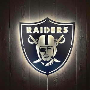 Op de afbeelding: Een zwart metalen wandbord in de vorm van een schild met het Raiders-logo, een centrale figuur met een ooglapje en twee gekruiste zwaarden. Het bord is verlicht met een helderwitte gloed, waardoor de uitgesneden tekst RAIDERS oplicht.