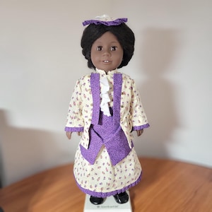 May include: A doll with dark skin and black hair, wearing a cream-colored dress with a floral pattern and purple accents. The doll is also wearing a matching jacket and hat. The doll is standing on a white base.