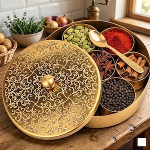 Könnte beinhalten: Eine runde Gewürzdose aus Messing mit einem aufwendig gravierten Blumendeckel und einem zentralen Knauf. Der offene Behälter enthält mehrere kreisförmige Fächer mit grünen Kardamomkapseln, rotem Chilipulver, Sternanis, Zimtstangen und schwarzen Pfefferkörnern.