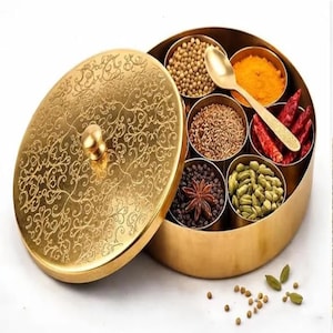 Könnte beinhalten: Eine runde Gewürzdose aus goldfarbenem Metall mit passendem Deckel, der eine filigrane Blumen-Gravur aufweist. Der Behälter ist in sieben Innenfächer unterteilt, die mit verschiedenen Gewürzen wie Kardamomkapseln, Chilischoten und einem goldenen Löffel gefüllt sind.