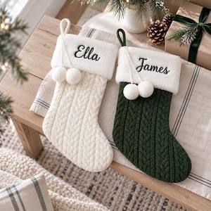 May include: Two cable-knit Christmas stockings with white faux fur cuffs. One stocking is cream with the name Ella embroidered in dark green, and the other is dark green with the name James embroidered in dark green. Both feature white pom-poms.