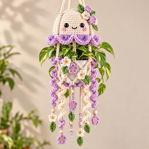 May include: A cream and purple crocheted jellyfish plant hanger featuring a smiling face with black eyes, pink cheeks, and a scalloped edge. The design includes dangling spiral tentacles, small crocheted flowers, green leaves, and wooden beads.