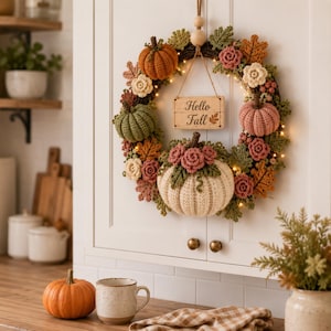 Può includere: Una ghirlanda circolare con zucche all'uncinetto in arancione, verde, rosa e crema, decorata con fiori e foglie all'uncinetto. Un cartello in legno appeso al centro mostra il testo Hello Fall. La ghirlanda è illuminata da piccole luci a corda.