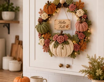 May include: A circular wreath featuring crocheted pumpkins in orange, green, pink, and cream, accented with crocheted flowers and leaves. A wooden sign hanging in the center displays the text Hello Fall. The wreath is illuminated by small string lights.