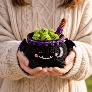 May include: A black crocheted cauldron with small purple wings, a purple scalloped rim, and a white crescent moon accent. The cauldron features a smiling face with pink cheeks and contains green crocheted spheres and a brown stirring stick.