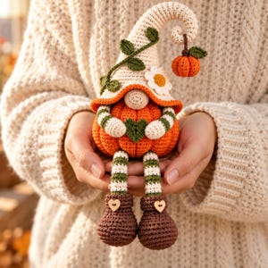 May include: A crochet gnome doll featuring a cream-colored hat with green vines and a white flower, a bright orange pumpkin body, and a small dangling pumpkin. The gnome has striped arms and legs, brown boots with heart buttons, and holds a green heart.