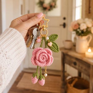 May include: A crochet keyring featuring a pink rose flower, two small pink buds, and two green leaves. The accessory includes a gold-toned star-shaped clasp, a small pink star charm, a white pearl bead, and a tiny pink heart charm attached to the ring.