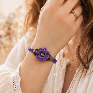 May include: A purple macrame flower bracelet featuring a central purple blossom with green leaves and small gold-toned beads at the center and sides. The bracelet is constructed from woven cord and secures around the wrist with a matching purple band.