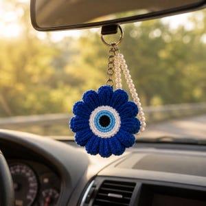 Puede incluir: Un adorno de coche de flor tejida a ganchillo con un diseño de ojo turco azul, blanco y negro en el centro. La flor está unida a un llavero de metal con una cadena corta y una hebra de perlas blancas, diseñado para colgar del espejo retrovisor.