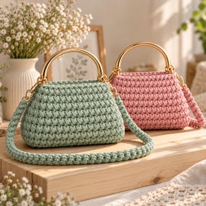 May include: Two crochet handbags, one in sage green and one in dusty pink, each featuring a structured gold-tone metal semi-circular handle and a matching thick crocheted shoulder strap attached with gold-tone metal swivel clasps.