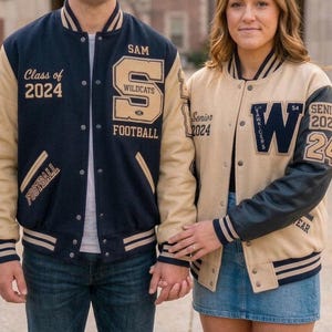 May include: Two varsity jackets. The left jacket is navy blue with tan sleeves, featuring 'SAM', 'Class of 2024', 'S', 'WILDCATS', and 'FOOTBALL' embroidery. The right jacket is tan with navy blue sleeves, featuring 'Senior 2024', 'W', and '24'.