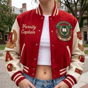 Puede incluir: Una chaqueta universitaria roja y crema con el texto Varsity Captain en el pecho, un escudo verde y dorado con el texto LIBERTIES ET SCIENTIA EST. 1881 y varios parches bordados en las mangas que incluyen letras, un casco y un balón de fútbol americano.