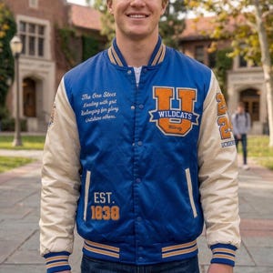 Op de afbeelding: Een blauw en crèmekleurig varsity-jack met oranje en wit borduurwerk. De voorkant bevat de tekst 'The One Stect Leading with grit Belonging for glory Outshine others', een 'U WILDCATS'-patch en 'EST. 1838'. De mouwen zijn crème met blauw en oranje gestreepte boorden.