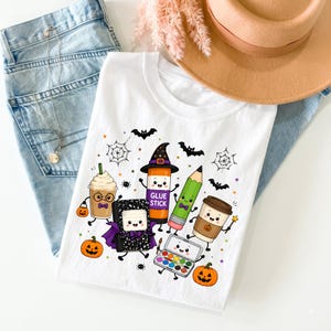 Puede incluir: Una camiseta blanca con un estampado gr&aacute;fico colorido de material escolar antropom&oacute;rfico, que incluye un pegamento en barra con sombrero de bruja, un l&aacute;piz, un cuaderno con capa, una caja de pinturas y dos tazas de caf&eacute;, rodeados de murci&eacute;lagos, ara&ntilde;as y calabazas.