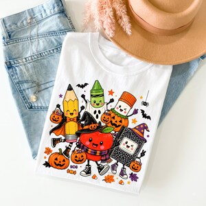 Puede incluir: Una camiseta blanca con un gr&aacute;fico colorido de &uacute;tiles escolares antropom&oacute;rficos disfrazados para Halloween, incluyendo un l&aacute;piz, cray&oacute;n, pegamento, manzana y cuaderno, rodeados de peque&ntilde;as calabazas, murci&eacute;lagos y hojas de oto&ntilde;o.