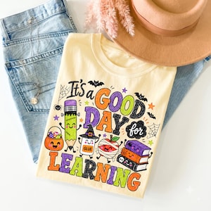 Puede incluir: Una camiseta de color amarillo p&aacute;lido con un gr&aacute;fico colorido que incluye el texto "It's a GOOD DAY for LEARNING" rodeado de ilustraciones de un l&aacute;piz, un pegamento, libros y una calabaza, decorado con murci&eacute;lagos, estrellas y telara&ntilde;as.