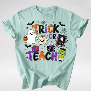 Puede incluir: Una camiseta de color verde menta claro con un gr&aacute;fico colorido de Halloween que incluye el texto "TRICK OR TEACH". El dise&ntilde;o muestra un fantasma con un cubo de calabaza, una manzana momia, dos regalos, un libro y un l&aacute;piz con cara.