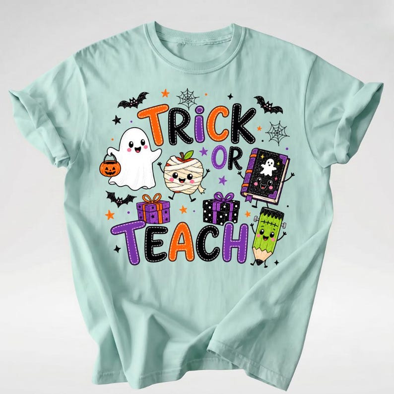 Puede incluir: Una camiseta de color verde menta claro con un gr&aacute;fico colorido de Halloween que incluye el texto "TRICK OR TEACH". El dise&ntilde;o muestra un fantasma con un cubo de calabaza, una manzana momia, dos regalos, un libro y un l&aacute;piz con cara.