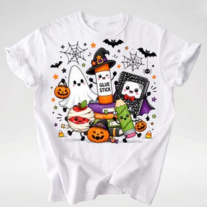 Puede incluir: Una camiseta blanca con un gr&aacute;fico colorido de Halloween que muestra un fantasma, un pegamento en barra con sombrero de bruja, una manzana momia, un cuaderno, un l&aacute;piz y una calabaza, todos con caras sonrientes, rodeados de murci&eacute;lagos.