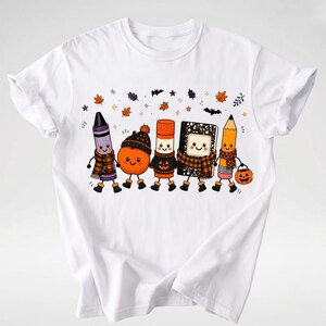 Puede incluir: Una camiseta blanca con una ilustraci&oacute;n gr&aacute;fica de cinco &uacute;tiles escolares antropom&oacute;rficos, incluyendo un cray&oacute;n morado, una calabaza naranja, un pegamento en barra, un cuaderno en blanco y negro y un l&aacute;piz amarillo, todos con bufandas.