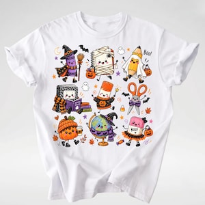Puede incluir: Una camiseta blanca con un estampado gr&aacute;fico colorido de varios &uacute;tiles escolares antropom&oacute;rficos disfrazados de Halloween, incluyendo un cray&oacute;n bruja, un cuaderno momia, un l&aacute;piz fantasma y un globo terr&aacute;queo con sombrero de bruja, con el texto Boo!