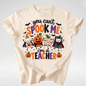 Puede incluir: Una camiseta de color crema con un gr&aacute;fico que incluye el texto "you can't SPOOK ME i'm a TEACHER". El dise&ntilde;o muestra personajes de dibujos animados: un bote de pegamento, una calabaza, una manzana momia y un libro, todos con capas.