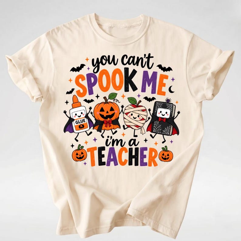Puede incluir: Una camiseta de color crema con un gr&aacute;fico que incluye el texto "you can't SPOOK ME i'm a TEACHER". El dise&ntilde;o muestra personajes de dibujos animados: un bote de pegamento, una calabaza, una manzana momia y un libro, todos con capas.