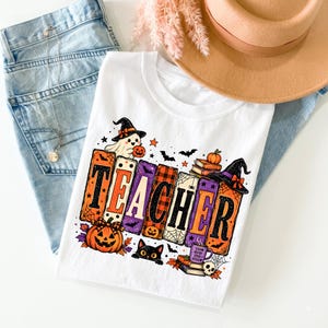 Puede incluir: Una camiseta blanca con un gr&aacute;fico que muestra la palabra TEACHER en letras grandes, coloridas y estampadas. El dise&ntilde;o incluye un fantasma con sombrero de bruja, una calabaza, un gato negro, libros, una telara&ntilde;a y una taza con el texto Teach Inspire Spook Repeat.