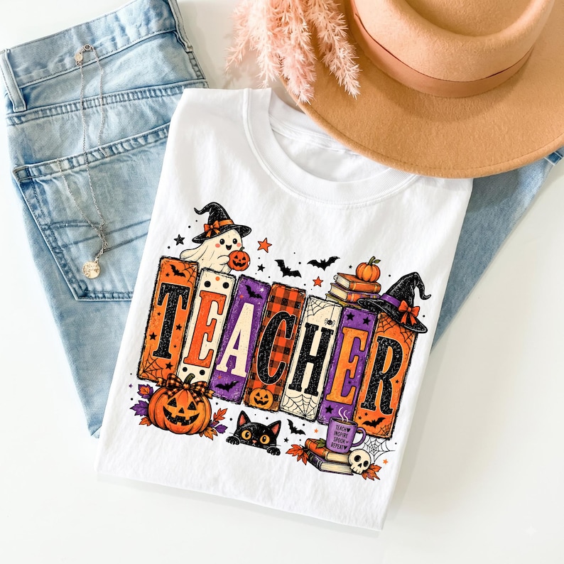Puede incluir: Una camiseta blanca con un gr&aacute;fico que muestra la palabra TEACHER en letras grandes, coloridas y estampadas. El dise&ntilde;o incluye un fantasma con sombrero de bruja, una calabaza, un gato negro, libros, una telara&ntilde;a y una taza con el texto Teach Inspire Spook Repeat.