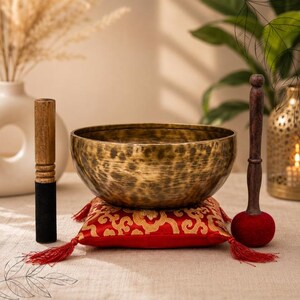 May include: A round, hammered brass singing bowl rests on a square red silk cushion with gold embroidered patterns and red tassels at each corner. A wooden mallet with a black felt tip and a wooden striker with a red felt ball are placed nearby.