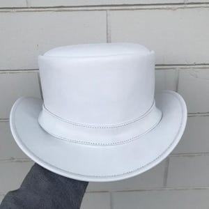 Op de afbeelding: Een effen witte hoge hoed met een platte kroon en een brede, gebogen rand. De hoed is voorzien van een gestructureerde band rond de basis met zichtbare stiksels langs de randen, wat zorgt voor een uniforme witte kleur over het hele ontwerp.