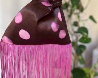 Può includere: Una borsa a nodo in raso marrone con un motivo a grandi pois rosa. Il bordo inferiore della borsa è rifinito con lunghe e fitte frange in una tonalità rosa vivace. Il manico è formato da un passante in tessuto annodato sulla parte superiore.
