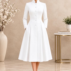 Puede incluir: Un vestido blanco de largo midi con cuello mao alto, mangas largas y cierre frontal de botones. La prenda presenta un corpiño ajustado con una falda acampanada y plisada que llega por debajo de las rodillas, incorporando bolsillos laterales.