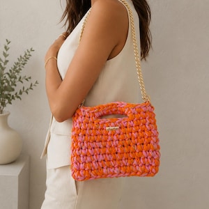 Può includere: Una borsa a mano rettangolare all'uncinetto caratterizzata da un mix vibrante di filato arancione neon e rosa acceso. La borsa include una tracolla a catena in metallo dorato e una piccola targhetta rettangolare con logo dorato sul davanti.